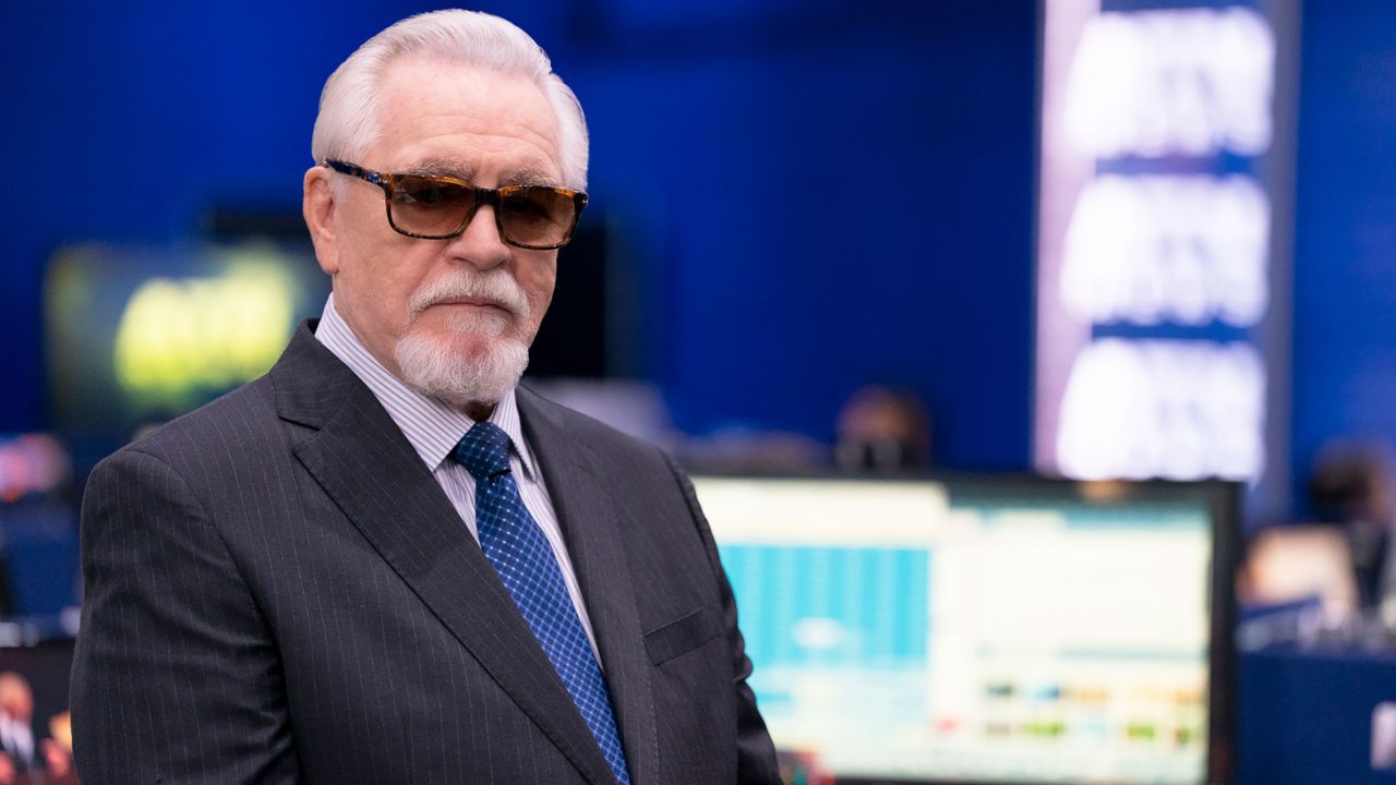 Brian Cox as logan roy on HBO's 'Succession.' He's an old, white man with white hair with a receding hairline, and a white goatee. He's wearing a black, pinstripe suit, with a purple-striped buttondown shirt and a blue tie with a white criss-cross pattern on it. He's wearing sunglasses while standing in a newsroom. We see him from the chest up.