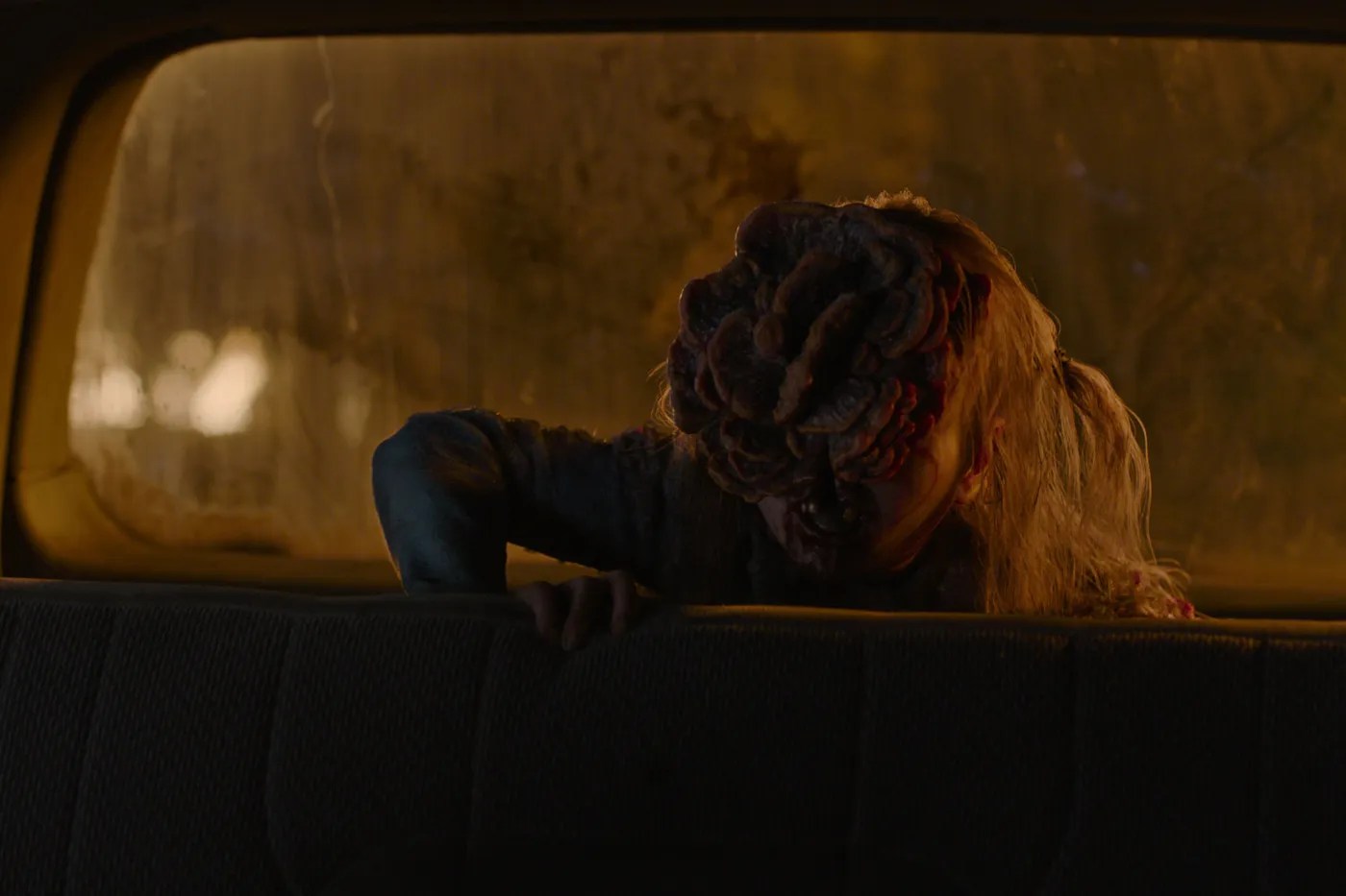 Image of a chiId clicker emerging from the back seat of a car at night on HBO's 'The last of Us.' She has blonde hair in a messy ponytail. The clicker cordyceps fungal plates are emerging out of her face obscuring everything but her mouth. 