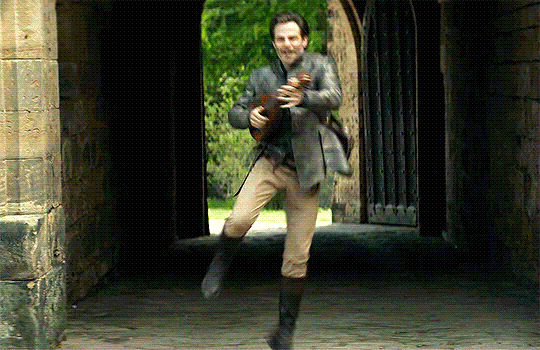 GIF of Chris Pine as Edgin in 'Dungeons & Dragons: Honor Among Thieves.' He is entering the courtyard of a castle while playing the lute and kicking up his heels. He's a white man with short, brown hair wearing a grey, leather jacket, khaki pants, knee-high black boots, and his lute case on his back. 