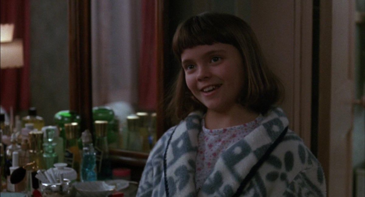 Christina Ricci as Kate Flax in 'Mermaids.' She's smiling as she stands in a bedroom with a vanity table behind her covered in perfume bottles. Her light brown hair is in a chin-length bob with bangs, and she's wearing flower-print pajamas under a grey and white robe.