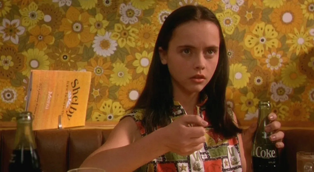 Image of Christina Ricci as young Roberta Martin in 'Now and Then.' She is looking skeptically at something as she holds up one hand in a fist and has a bottle of Coke in the other. She's wearing a colorful, sleeveless shirt, and her long hair is parted down the middle. She's sitting in a diner booth, and there's really loud, yellow-orange, flower-print wallpaper behind her.