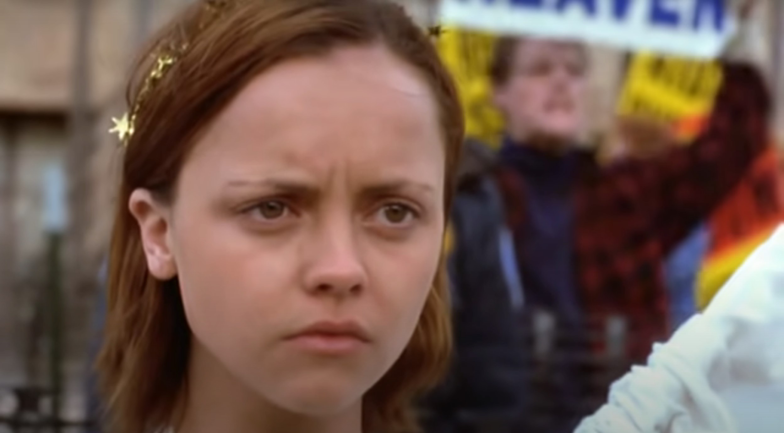Christina Ricci in 'The Laramie Project' (HBO). It's a close-up of her face, looking concerned. She's wearing a small star garland in her short, light-brown hair. There are angry protesters in the background.