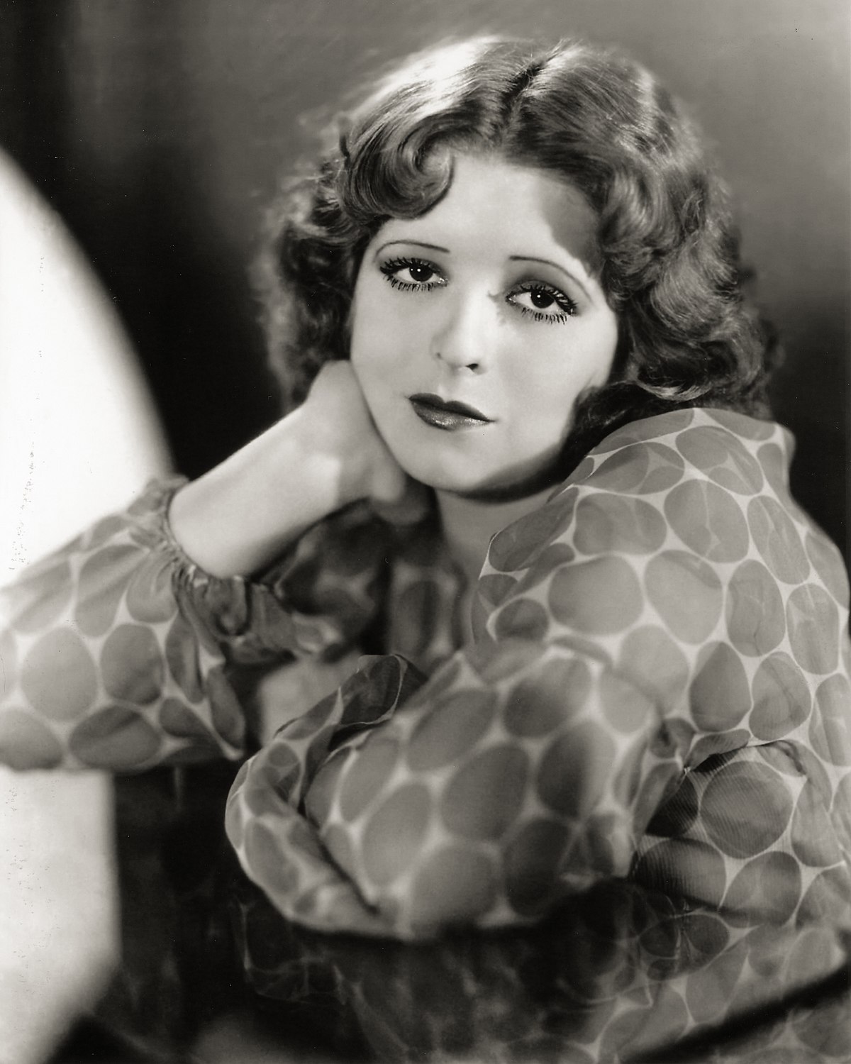 Image of actress, Clara Bow. It's a black and white headshot. Clara is a white woman with wavy, shoulder-length hair. She's wearing a gauzy, long-sleeved, polka-dot blouse, and she's wearing dark lipstick and thick mascara in the 1920s style. She's visible from the waist up, and she's straddling a chair sitting forward to lean against the back. One arm is across the chair back, the other holds up her chin. 
