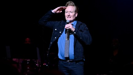 Conan O'Brien salutes, standing alone on a stage holding a microphone.