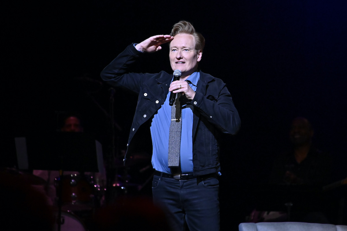 Conan O'Brien salutes, standing alone on a stage holding a microphone.