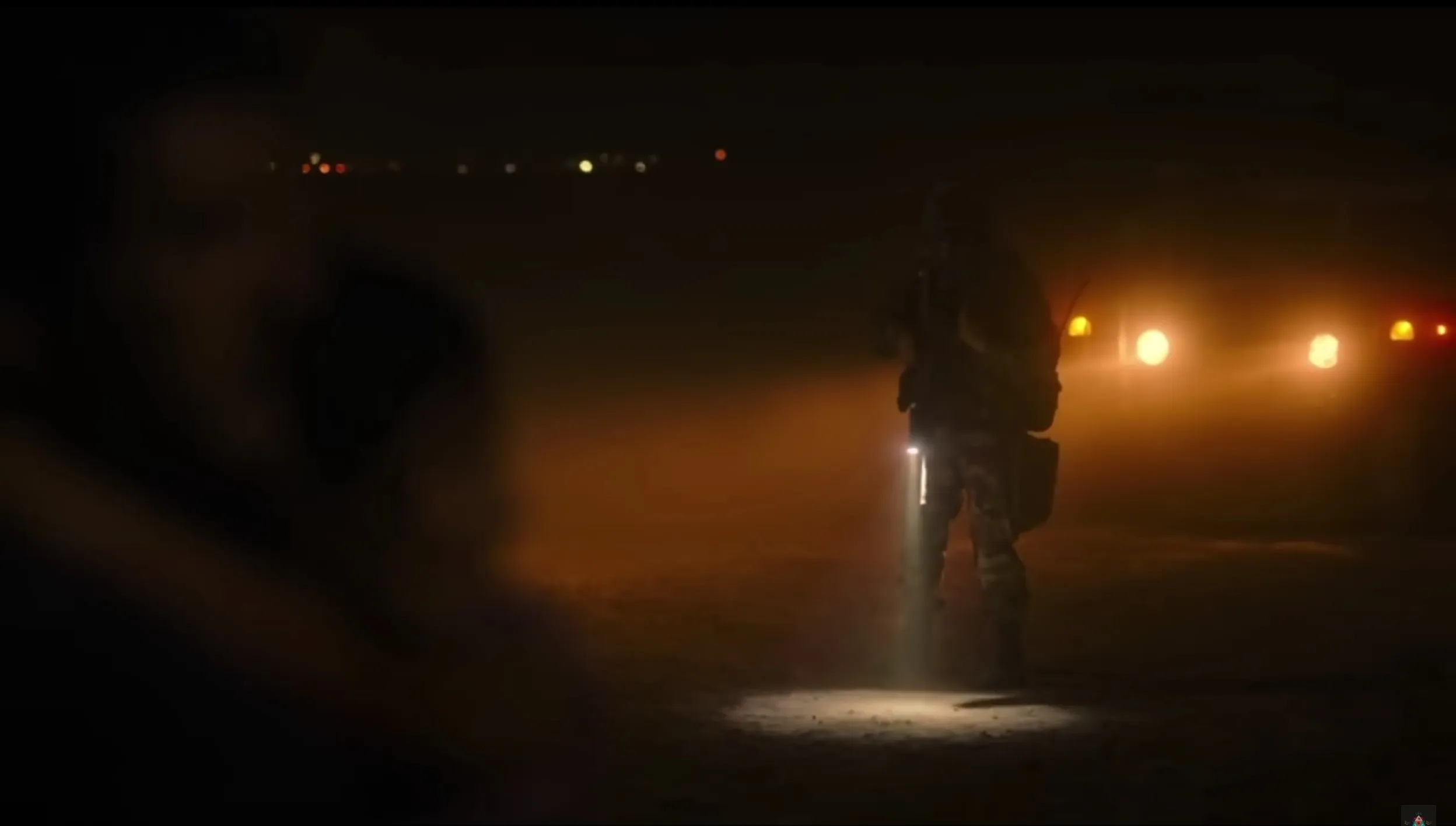 Screenshot of Craig Mazin in his uncredited role as a soldier in HBO's 'The Last of Us." The soldier is standing outside in the dark night, illuminated only by the headlights of a truck, and his own flashlight. Out of focus in the foreground, we see the silhouette of Joel (Pedro Pascal)  carrying Sarah (Nico Parker). 