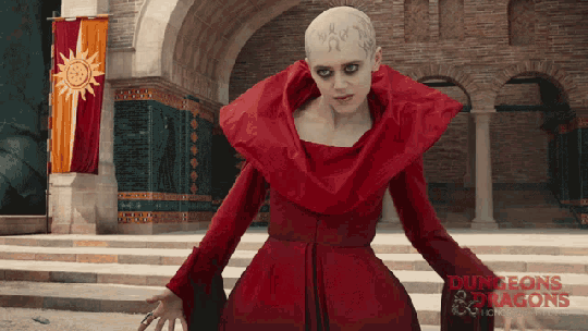 GIF of Daisy Head as Sofina in 'Dungeons & Dragons: Honor Among Thieves.' She is bald with pasty white skin and tattoos on her head. She's wearing a long, red dress with long sleeves and a high, wide collar. She shoots a spell out of her hand, forcefully throwing Edgin to the ground. 