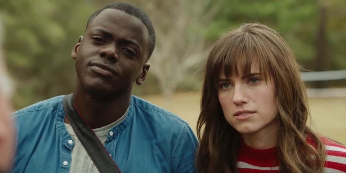 Image of Daniel Kaluuya (Chris) and Allison Williams (Rose) in 'Get Out' We see them from the shoulders up standing outdoors side by side. Chris is on the left wearing a blue jacket and a grey shirt. A camera strap hangs across his body. Rose has long, brown hair with bangs. She's wearing a red and grey striped shirt. 