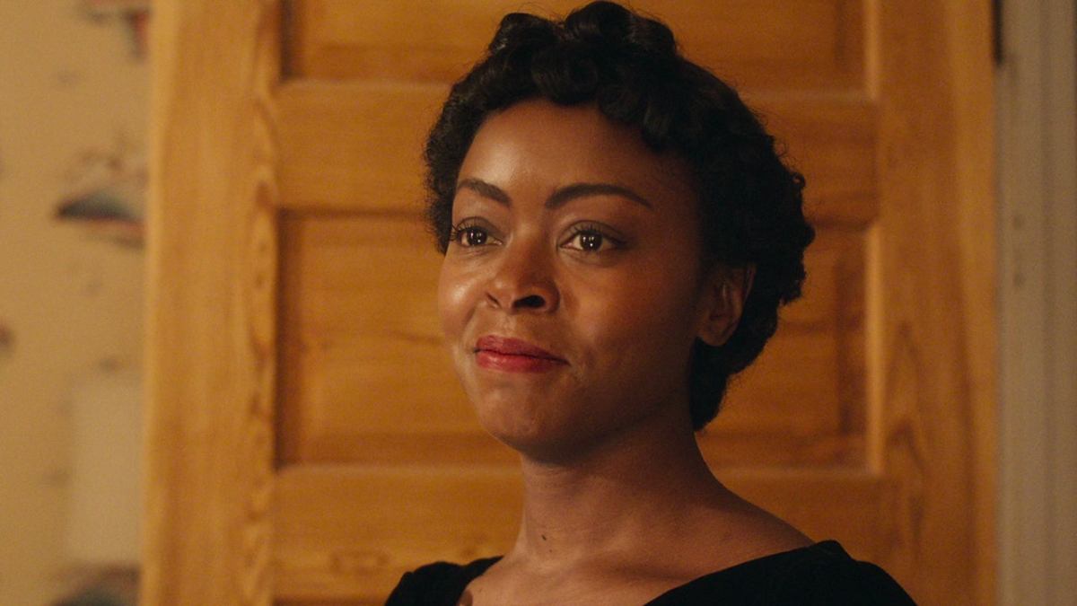 Danielle Deadwyler in a scene from 'Till.' She is indoors, standing in front of an open wooden bedroom door. We see her from the shoulders up. She's wearing a black dress and wears red lipstick. She has a soft smile on her face, and her eyes look teary.