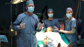 Image of Darren Dolynski (Surgeon), Bella Ramsey (Ellie), Ana Rice (Nurse), and Laura Bailey (Nurse) in HBO's 'The Last of Us.' The surgeon and nurses are are all wearing blue scrubs, white surgical gloves, blue surgical masks, and blue head coverings as they surround Ellie, who is unconscious on a gurney. The surgeon is in the foreground on the left, wearing clear, plastic goggles, and all of them are looking at something in the direction of the camera.