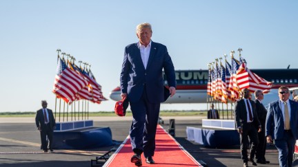 Donald Trump walks down a long red carpet outside with flags and his campaign sign in the background.