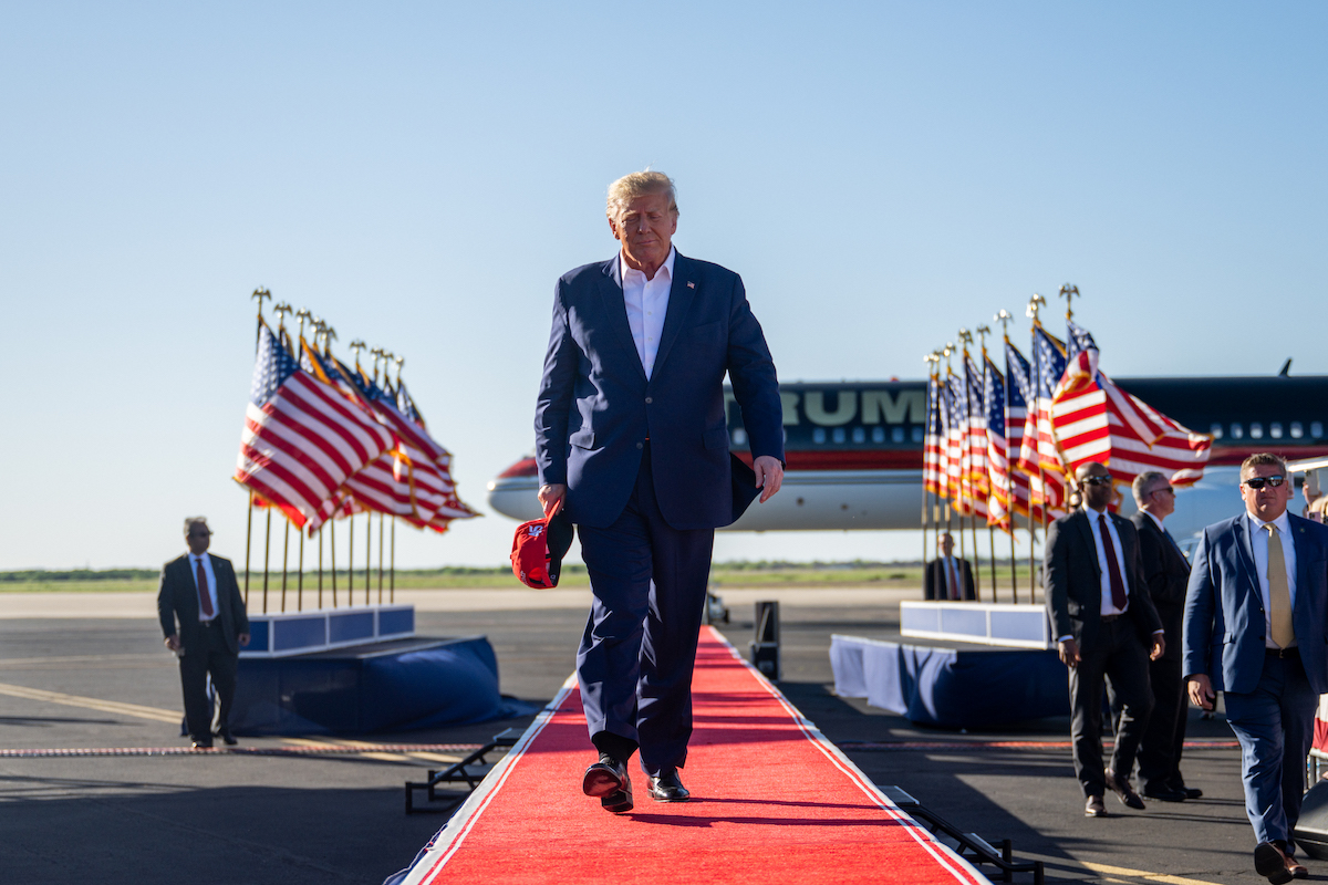 Donald Trump walks down a long red carpet outside with flags and his campaign sign in the background.