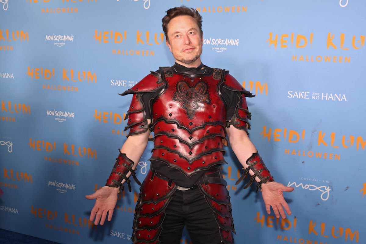 Elon Musk in a costume of red armor, poses and shrugs on a red carpet.