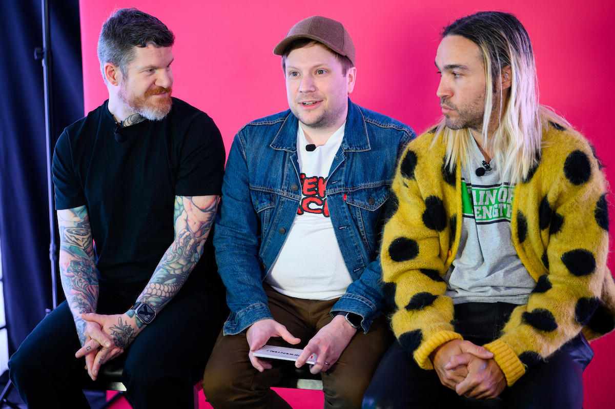 Three men (the members of Fall Out Boy) sit next to each other during an interview, in front of a pink screen.
