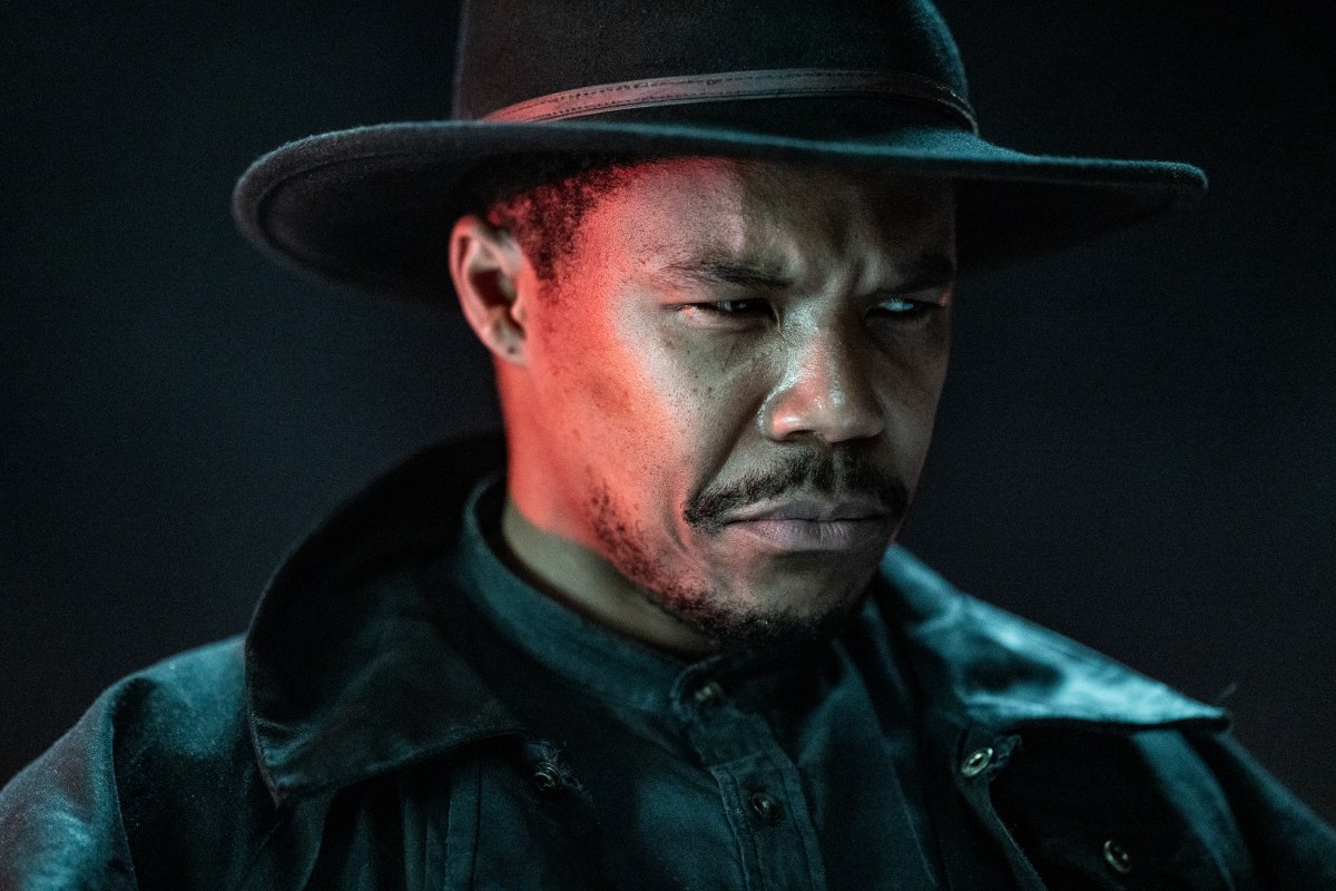 Publicity image of Gaius Charles as Perlie Armstrong on AMC's 'The Walking Dead: Dead City.' We see him from the shoulders up. He is a Black man wearing a wide-brimmed fedora/cowboy hat, a black leather jacket, and a denim button-down shirt. He has a mustache and the beginnings of a beard. He is scowling sternly and looking off to the side.