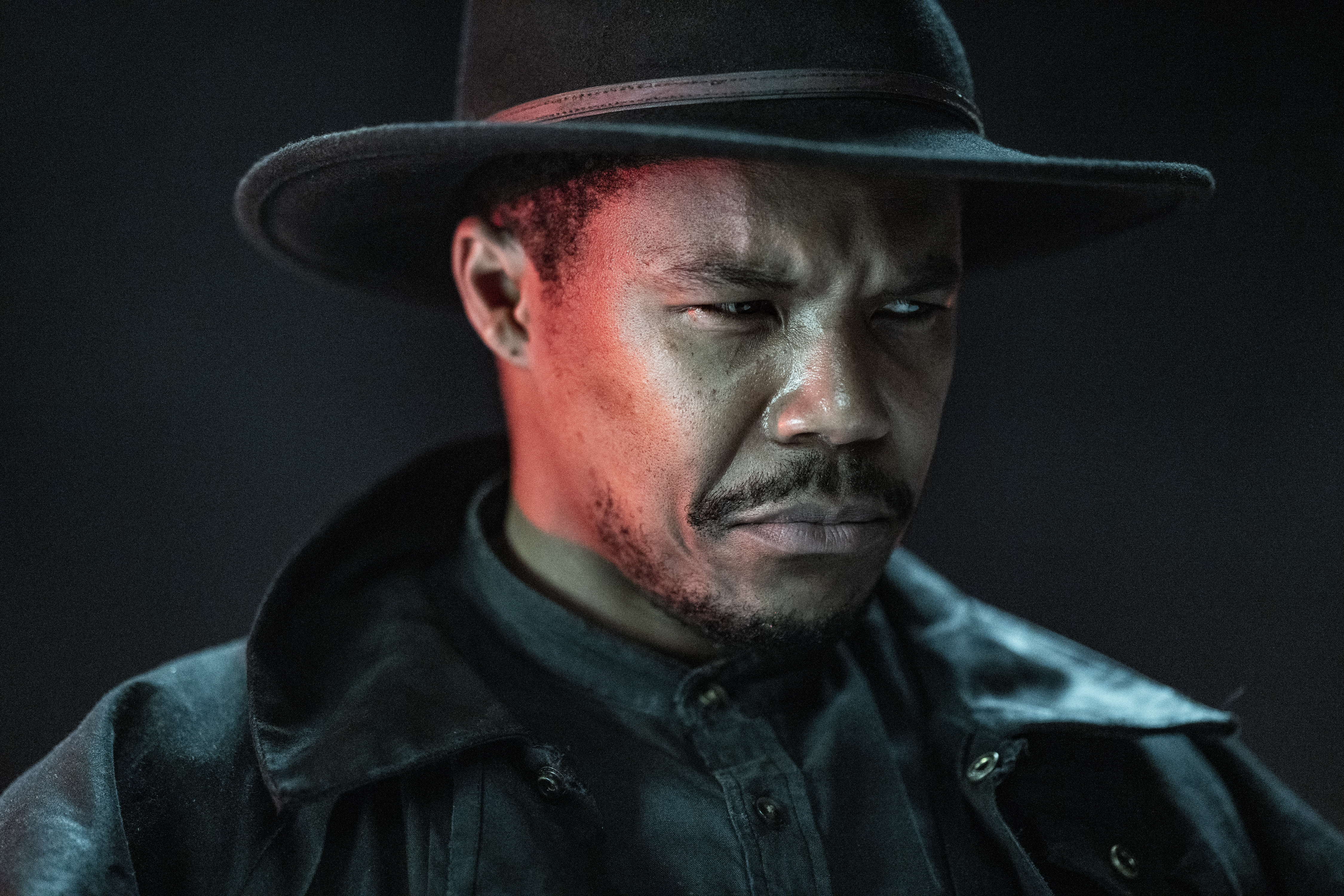 Publicity image of Gaius Charles as Perlie Armstrong on AMC's 'The Walking Dead: Dead City.' We see him from the shoulders up. He is a Black man wearing a wide-brimmed fedora/cowboy hat, a black leather jacket, and a denim button-down shirt. He has a mustache and the beginnings of a beard. He is scowling sternly and looking off to the side. 