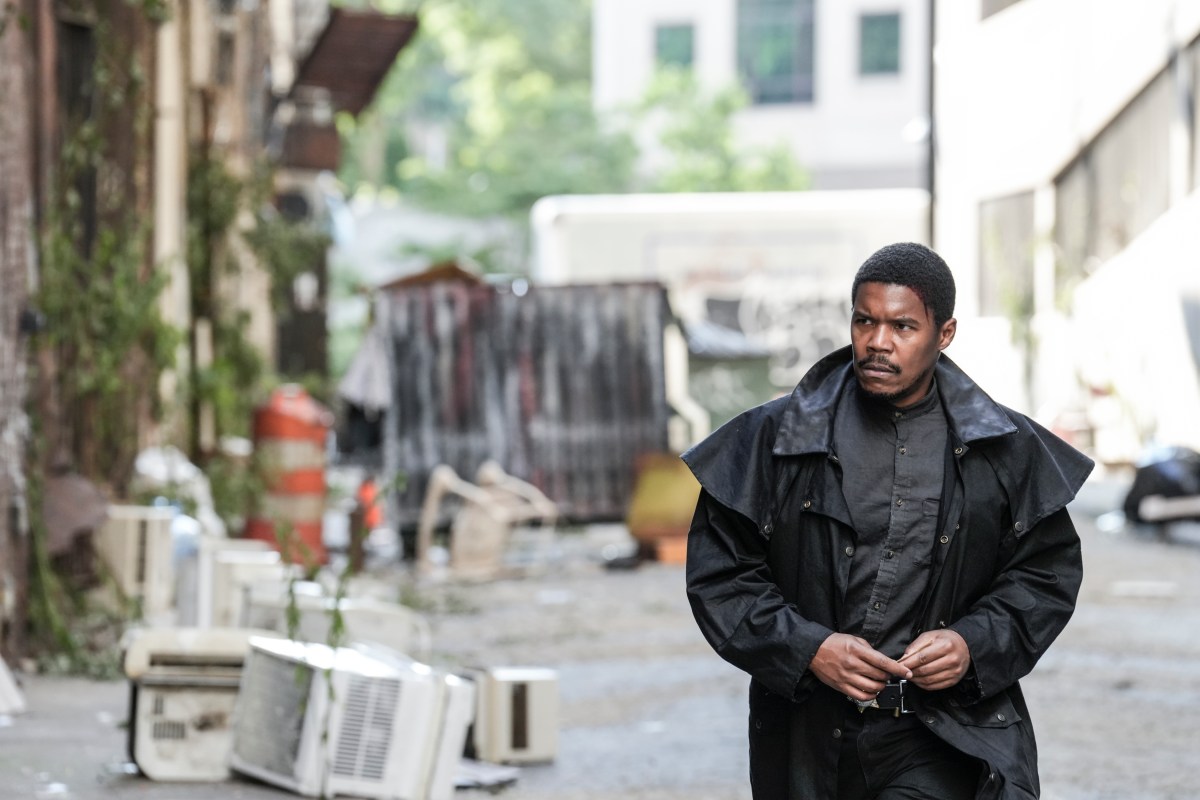 Image of Gaius Charles as Perlie Armstrong on AMC's 'The Walking Dead: Dead City.' We see him from the waist up. He is a Black man wearing a black jacket that looks like a cloak, and a grey button-down shirt. He's walking down an alley and holding a corner of his jacket with both hands. He has close-cropped hair and a mustache. He's looking sternly off to the side.
