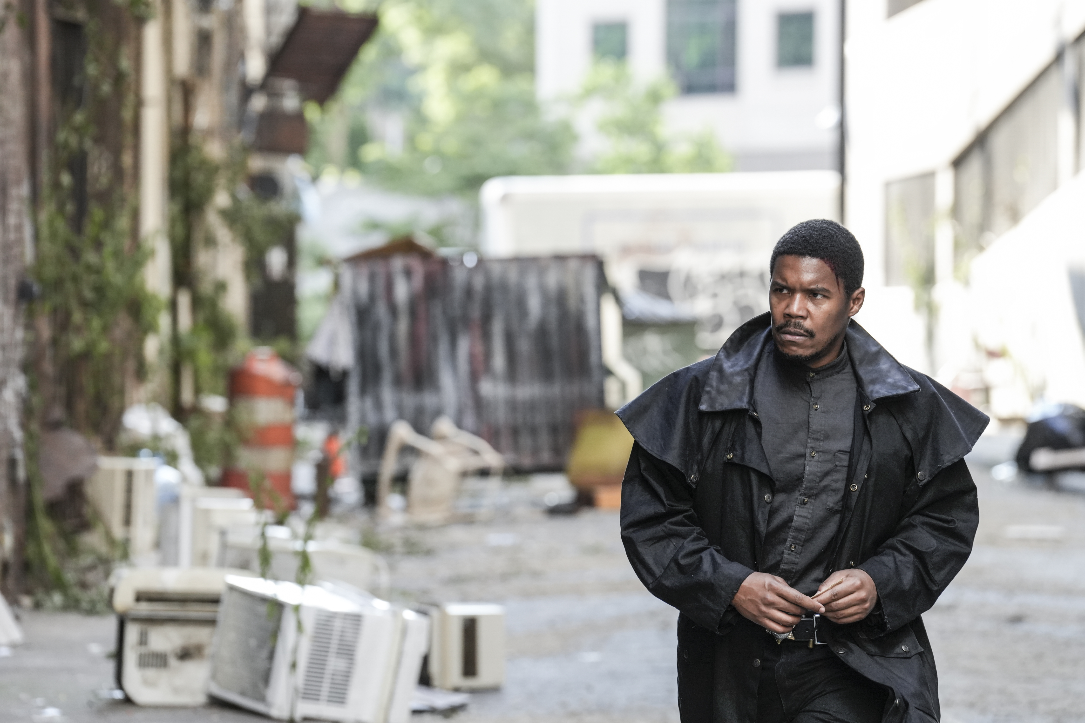 Image of Gaius Charles as Perlie Armstrong on AMC's 'The Walking Dead: Dead City.' We see him from the waist up. He is a Black man wearing a black jacket that looks like a cloak, and a grey button-down shirt. He's walking down an alley and holding a corner of his jacket with both hands. He has close-cropped hair and a mustache. He's looking sternly off to the side. 