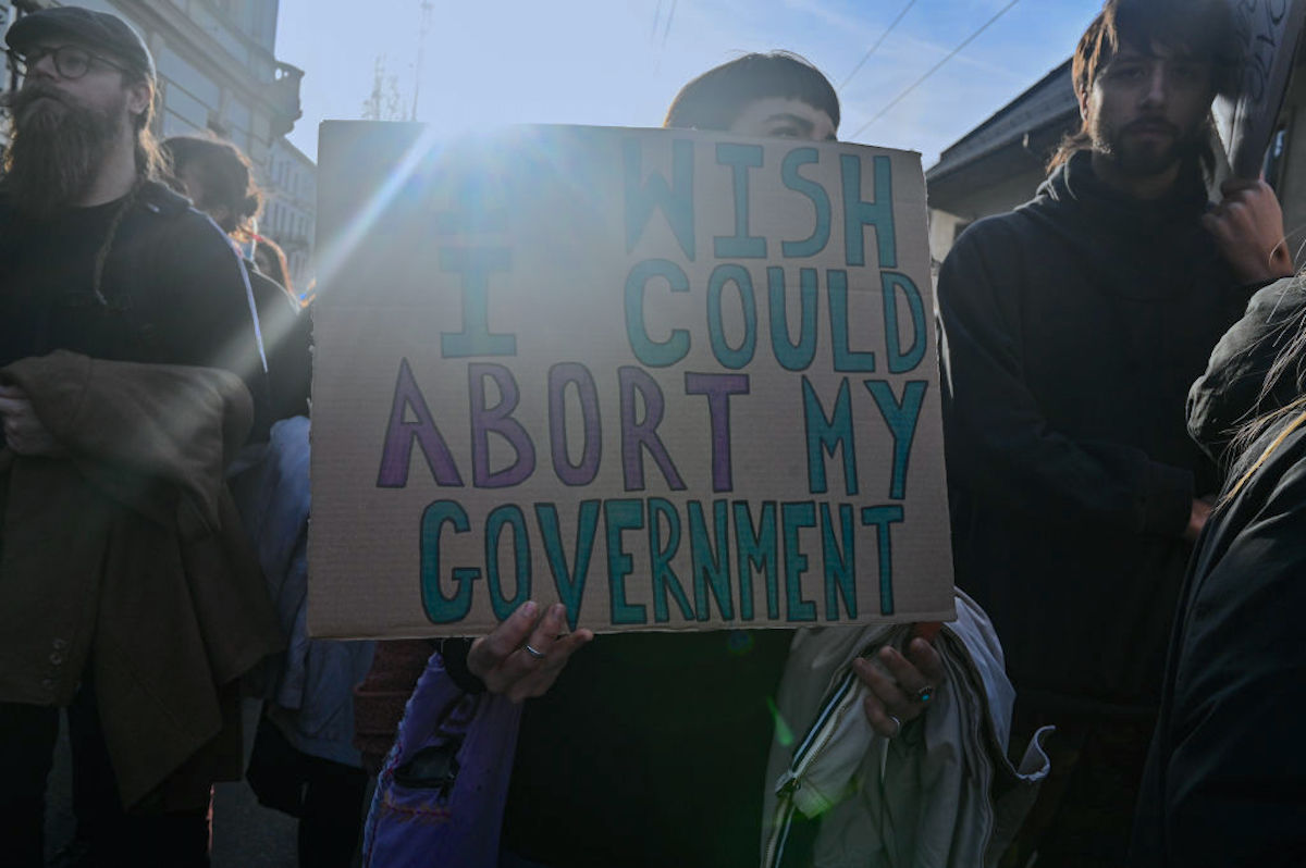 A woman at a protest holds a cardboard sign reading "I wish I could abort my government"