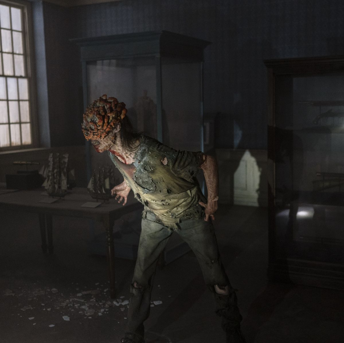 Full-length image of an adult clicker in a museum on HBO's 'the last of us.' It's body is turned toward the left as if it hears something in that direction. Thick fungal plates cover most of the top of its head, though some long dark hair is visible, too. It's wearing a torn green shirt and dark torn pants. 