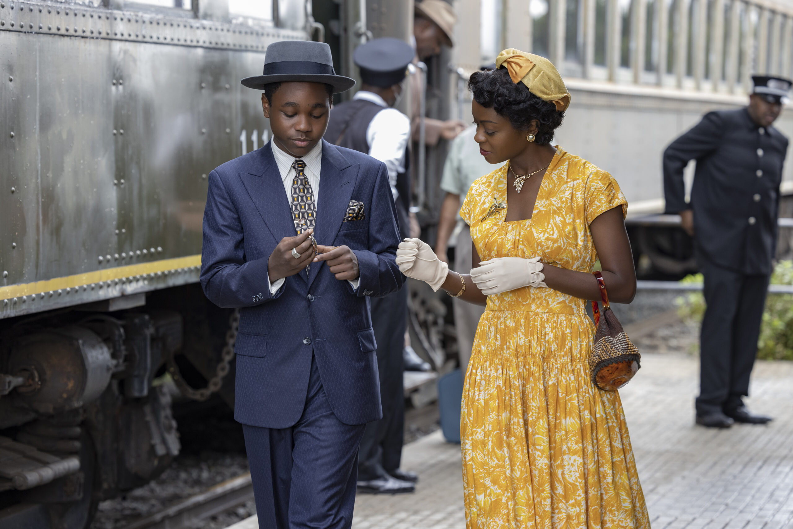 Jalyn Hall as Emmett Till and Danielle Deadwyler as Mamie Till Bradley in a scene from 'TILL.' They are standing on a train platform next to a train that's pulled into the station. Emmett is in a dark blue suit and hat looking down at a watch in his hands as he walks. Mamie is in a bright yellow and white floral print dress and a yellow hat. She has short hair that's set in waves. She's looking at what her son is doing with the watch. She's wearing white gloves and has a small purse hanging from one arm. There are train employees in the background.