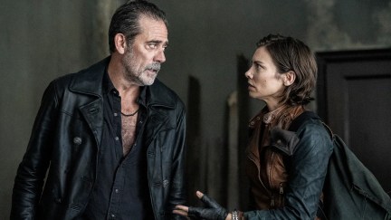 Jeffrey Dean Morgan as Negan and Lauren Cohan as Maggie in a scene from AMC's 'The Walking Dead: Dead City.' Negan (left) is a tall, white man with slicked-back black hair with a receding hairline and a salt-and-pepper beard. He's wearing all black - a black leather jacket, a black button-down with the buttons open to mid-chest, and a thin, black necklace. He's looking to the side at Maggie. Maggie (right) is in profile looking at him. She's a white woman with chin-length brown hair, and she's wearing a long-sleeved, black shirt underneath a brown, leather vest. She has a black backpack over her shoulder.