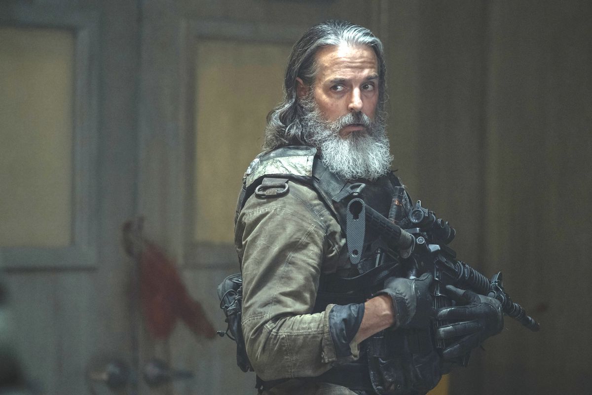 Image of Jeffrey Pierce as Perry in HBO's 'The Last of Us.' He's standing in a room with a set of double doors behind him. We see him from the waist up. He's got shoulder-length salt-and-pepper hair and a grey beard. He's wearing green military fatigues and is carrying a machine gun close to his chest. He's turned to the side looking at something approaching. 