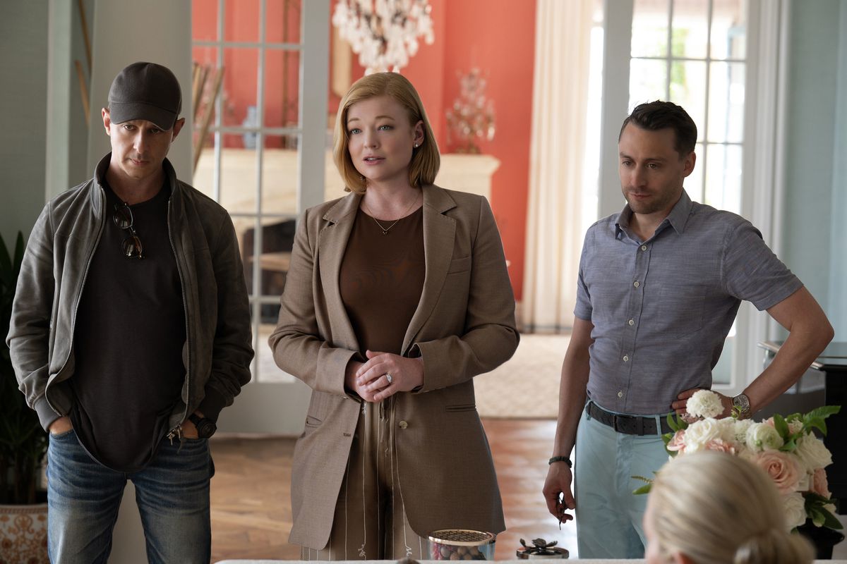 Image of Jeremy Strong as Kendall, Sarah Snook as Sibohan, and Kieran Culkin as Roman on HBO's 'Succession.' They are standing in an upscale home. Kendall has his hands in his pockets, and is wearing a black baseball cap, a grey fleece jacket over a black shirt, and jeans. Shiv's chin-length, red hair is parted on the side, and she's wearing a light brown blazer over a dark brown shirt and light brown linen pants. Her hands are clasped together, and she wears a large wedding ring. Roman is wearing a short-sleeved, periwinkle buttondown with the collar button open, and light blue pants with a dark belt. He has one hand on his hip.