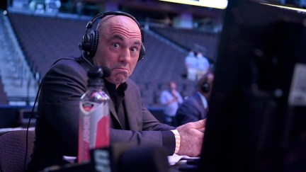 Joe Rogan mugs at the camera while sitting at a computer inside an arena, wearing headphones.