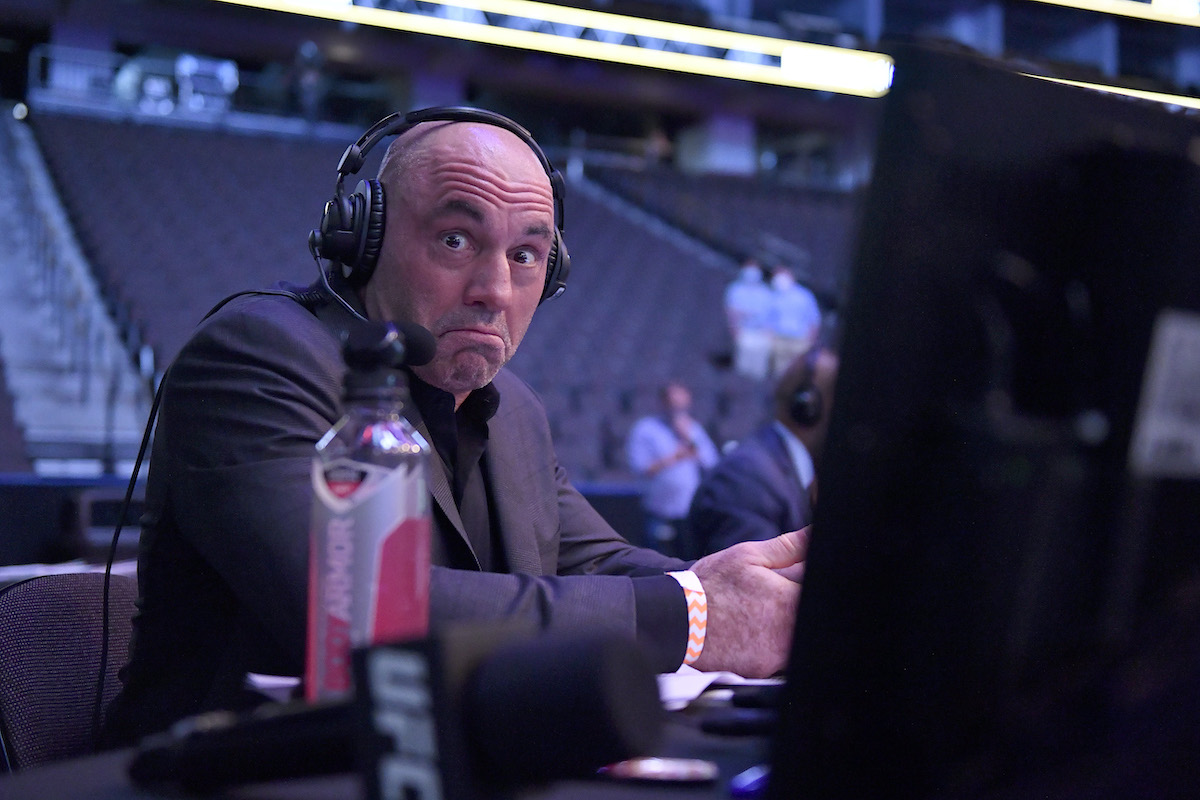 Joe Rogan mugs at the camera while sitting at a computer inside an arena, wearing headphones.