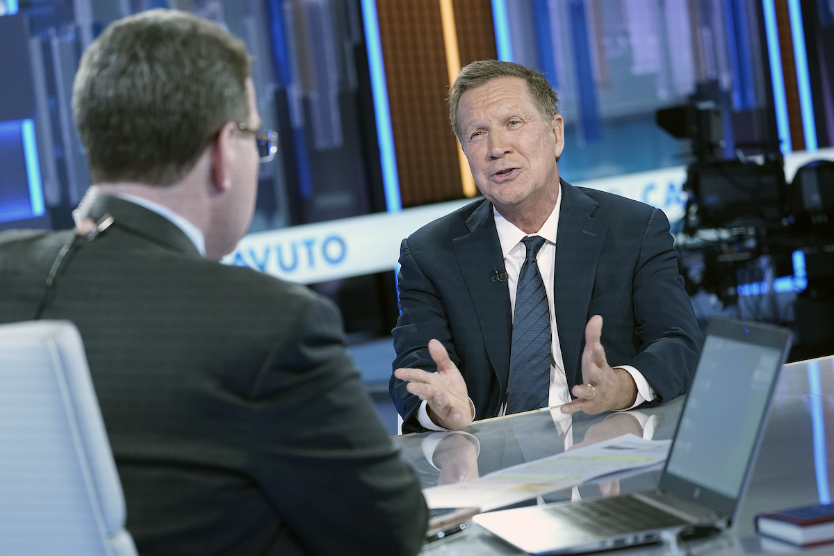 An older white man (Kasich) sits and speaks from a news desk, opposite another man.