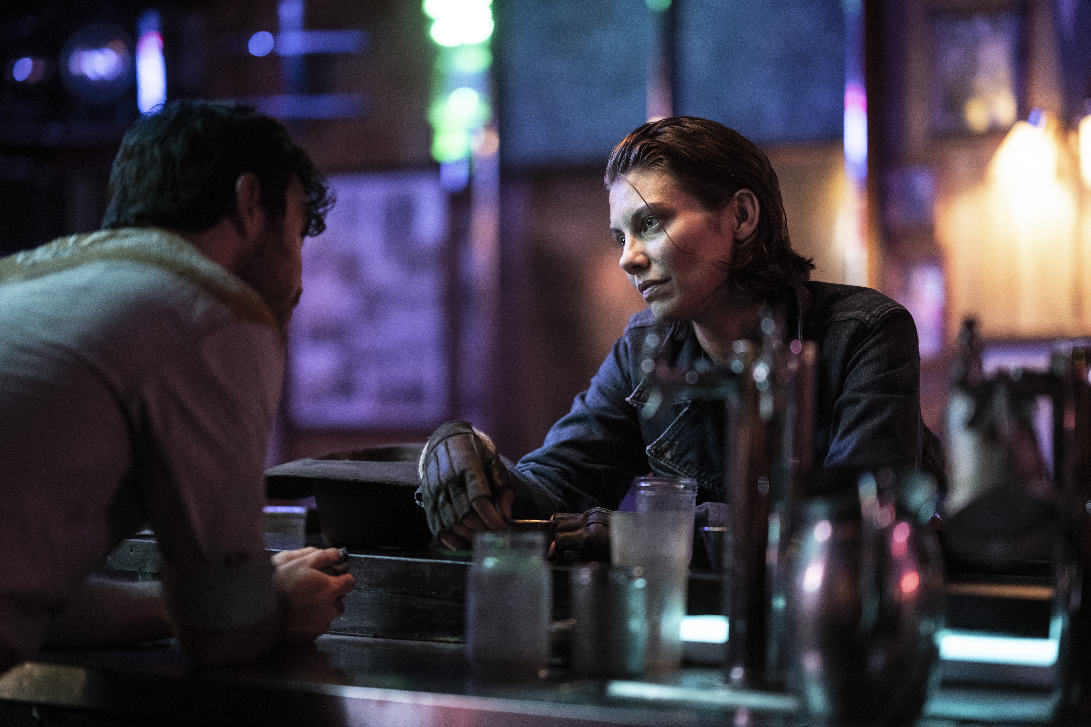 Image of Lauren Cohan as Maggie and Charlie Solis as Bartender in a scene from AMC's 'The Walking Dead: Dead City.' The Bartender, a white man with short, black hair and a beard and wearing a white, 3/4 sleeved button-down shirt has his back to the camera as he leans on the bar. Maggie is a white woman who is facing him as she sits at the bar, her short, chin-length brown hair slicked back with a tendril falling in her face. She's wearing a dark, long-sleeved shirt under a brown, leather vest, and black, fingerless gloves. She's listening intently to the Bartender as he speaks. 