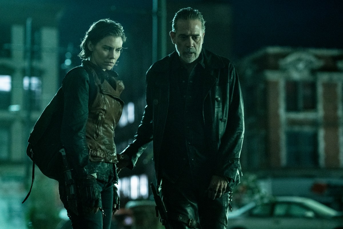 Lauren Cohan as Maggie and Jeffrey Dean Morgan as Negan in a scene from AMC's 'The Walking Dead: Dead City.' They are both looking down at something on the ground and are visible from the thighs up. Maggie (left) is a white woman with chin-length brown hair, and she's wearing a long-sleeved, black shirt underneath a brown, leather vest and black pants and fingerless gloves. She has a black backpack over her shoulder. Negan (right) is a tall, white man with slicked-back black hair with a receding hairline and a salt-and-pepper beard. He's wearing all black - a black leather jacket, a black button-down.