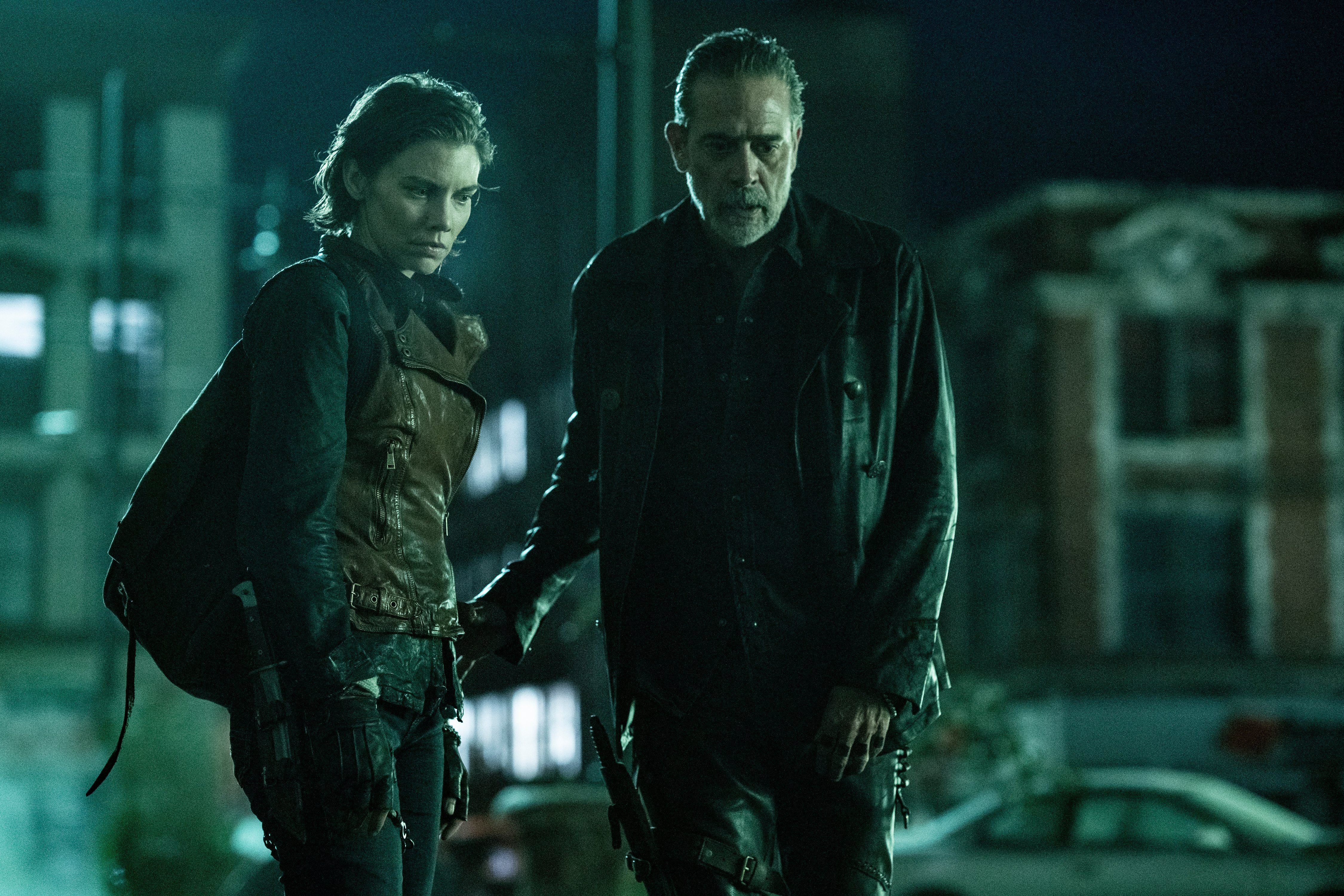 Lauren Cohan as Maggie and Jeffrey Dean Morgan as Negan in a scene from AMC's 'The Walking Dead: Dead City.' They are both looking down at something on the ground and are visible from the thighs up. Maggie (left) is a white woman with chin-length brown hair, and she's wearing a long-sleeved, black shirt underneath a brown, leather vest and black pants and fingerless gloves. She has a black backpack over her shoulder. Negan (right) is a tall, white man with slicked-back black hair with a receding hairline and a salt-and-pepper beard. He's wearing all black - a black leather jacket, a black button-down. 