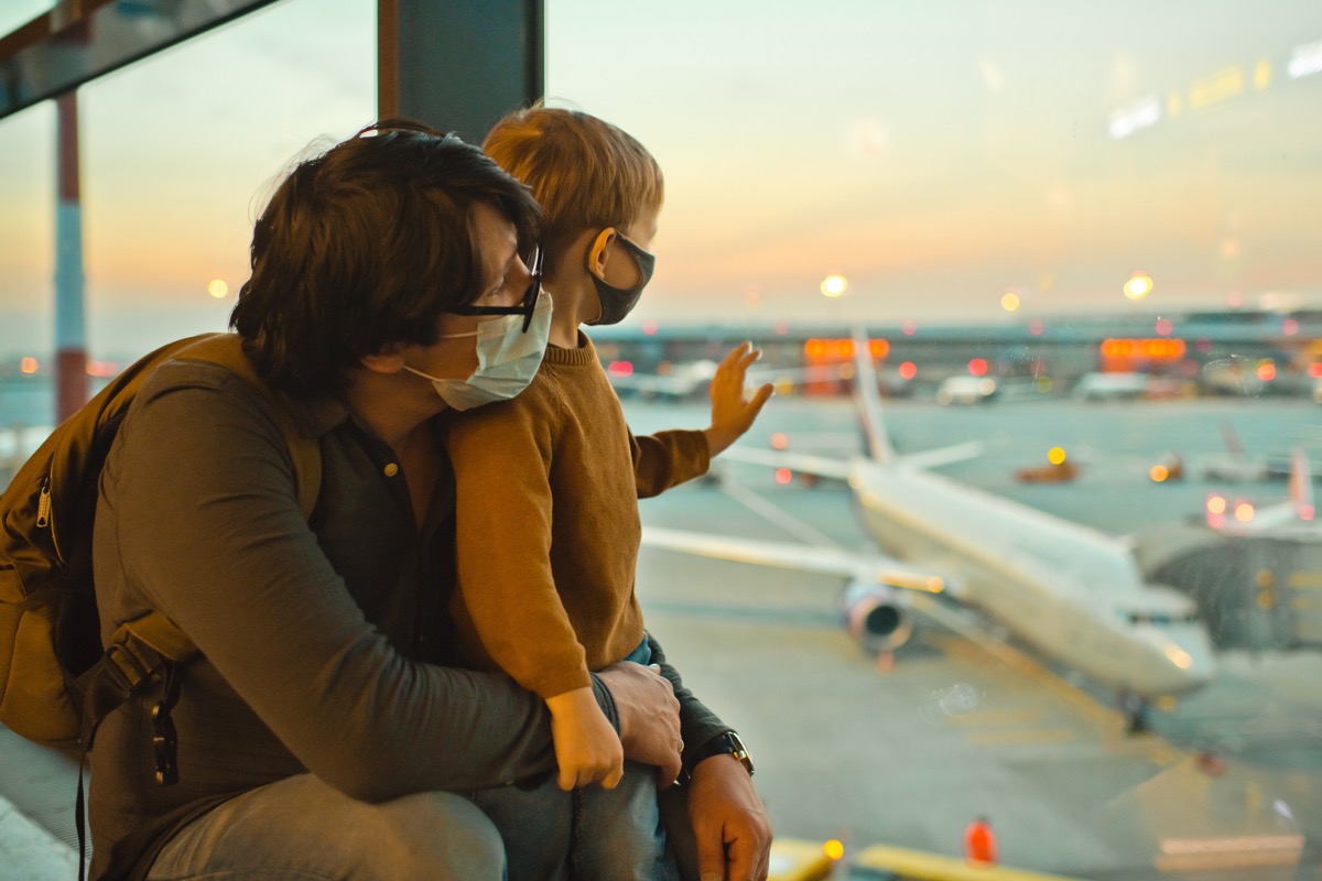 A father and son, wearing masks, watch the airplanes and wave from an airport window.