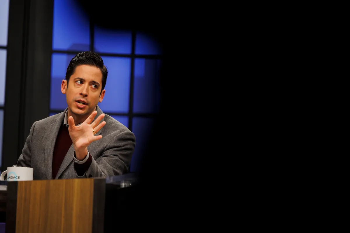 A man (Michael Knowles) waves his hand and looks irritated sitting at a TV news desk.