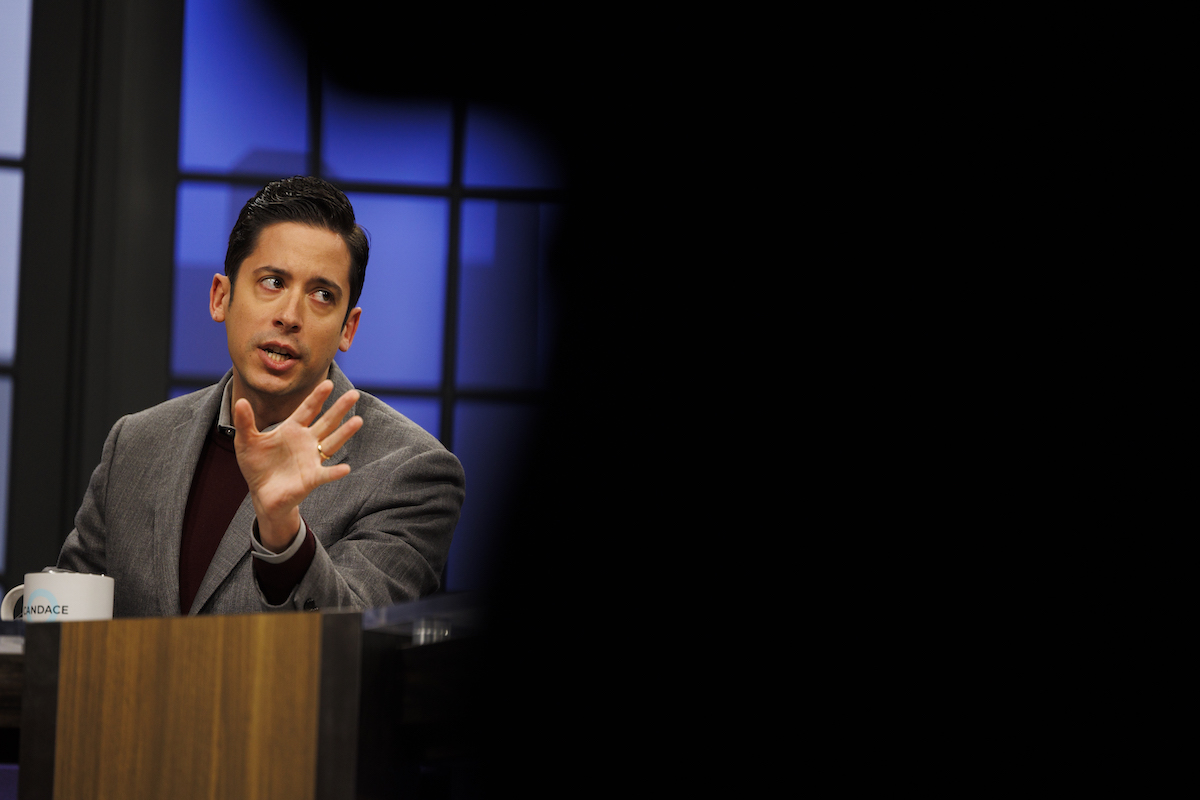 A man (Michael Knowles) waves his hand and looks irritated sitting at a TV news desk.
