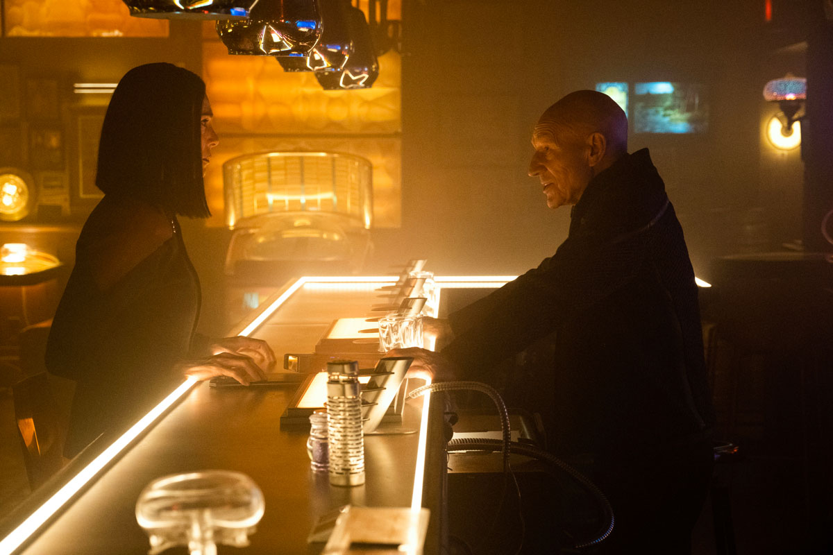 Image of Michelle Forbes as Ro Laren and Patrick Stewart as Jean-Luc Picard in a scene from "Star Trek: Picard." They are each standing on opposite sides of a bar. Picard in the bartender spot, Ro on the customer side. Ro's dark, shoulder-length hair obscures most of her face, which we see in profile. She has her hands on the bar and is wearing a black Starfleet uniform with dark red on the shoulders. Picard is bald and is wearing all black. His hands are also on the bar and we see him in profile angrily talking to Ro.