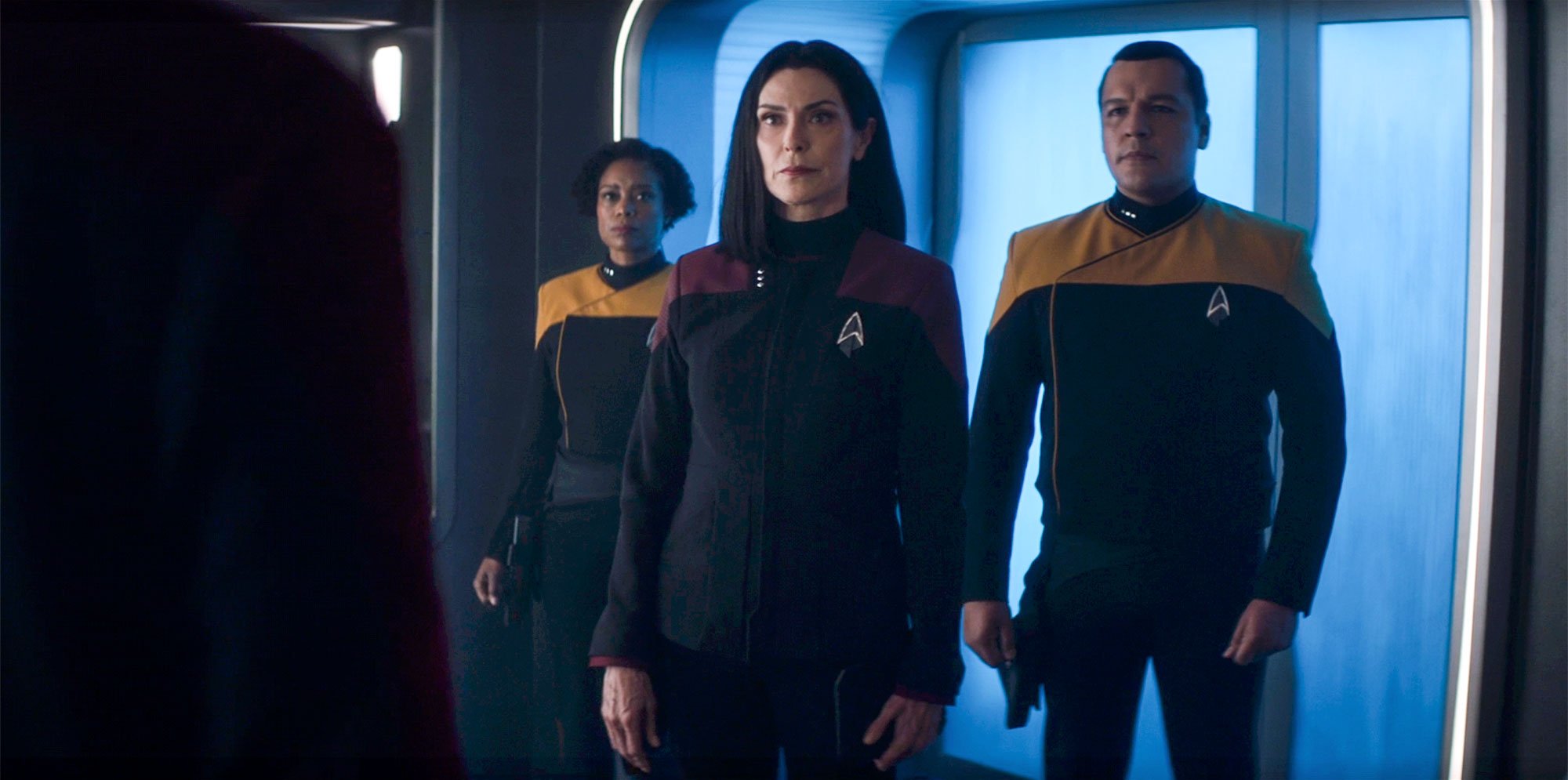 Image of Michelle Forbes as Ro Laren on 'Star Trek: Picard.' She's standing in a corridor on the Titan flanked by two Starfleet security officers. They're visible from the thighs up. Ro's dark, shoulder-length hair is parted down the middle. She's wearing a black Starfleet uniform with dark red shoulders. The security officer on the left is a Black woman with short, curly, dark hair who's wearing a black Starfleet uniform with gold shoulders. The security officer on the right is a man with brown skin in the same uniform as the woman on the left. The three of them are looking at someone standing in the corner of the frame out-of-focus.