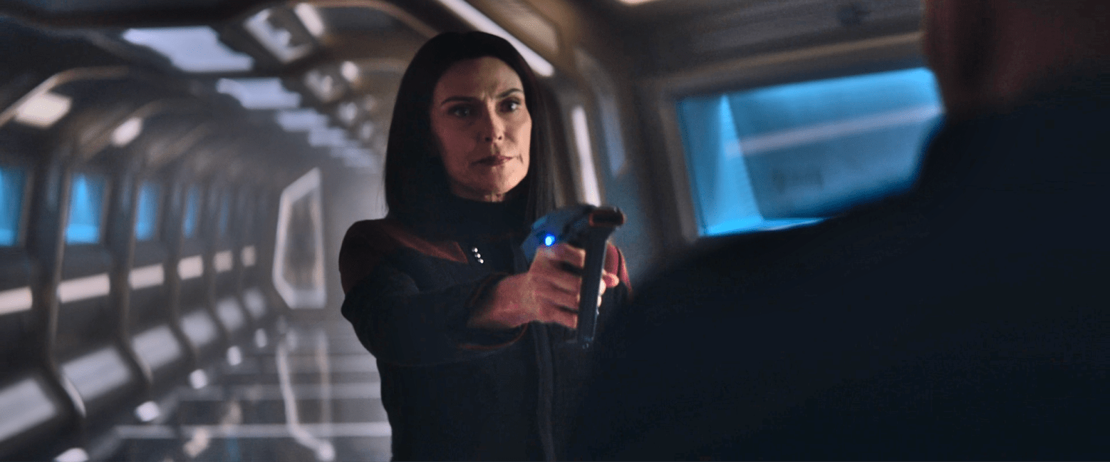 Image of Michelle Forbes as Ro Laren on 'Star Trek: Picard.' She stands in a ship corridor pointing a phaser at someone. Her shoulder-length dark hair is parted in the middle. She's wearing a black Starfleet uniform with dark red shoulders.