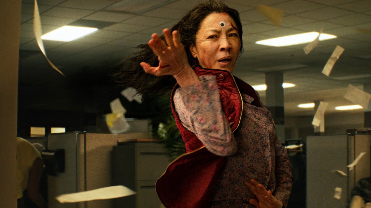 Image of Michelle Yeoh as Evelyn in 'Everything, Everywhere, All at Once.' She stands in a martial arts stance with her arms drawn back, ready to fight. She's in an office, and wind is blowing around her causing her long dark hair and maroon, quilted vest to be blowing around as well as papers everywhere. She has a googly eye stuck on her forehead between her eyes. 