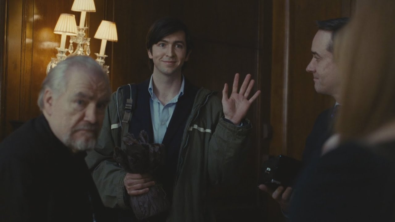 Image of Nicholas Braun as Greg Hirsch on HBO's 'Succession.' He is standing in a door at logan's apartment waving at the rest of the family. He's a tall, young, white man with short, shaggy hair and side bangs. He's wearing a green jacket over a dark blazer and a light blue buttondown with the collar button open. He's holding a bundle with one hand and waving with the other. He's surrounded by other family members who are slightly out of focus in the shot.