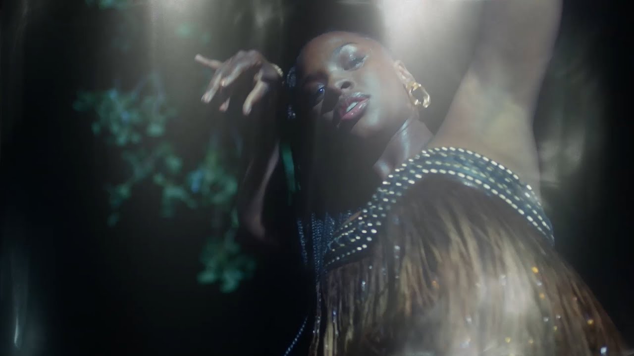 Image of Nirine S. Brown as Ni'jah in Amazon's 'Swarm.' We see Ni'jah through a haze in a scene from a music video. She is a Black woman with her hair in a long ponytail, gold hoop earrings, and wearing a dress made of brown and green flapper-type dress. Her arms are over her head as she dances, looking into the camera from above.