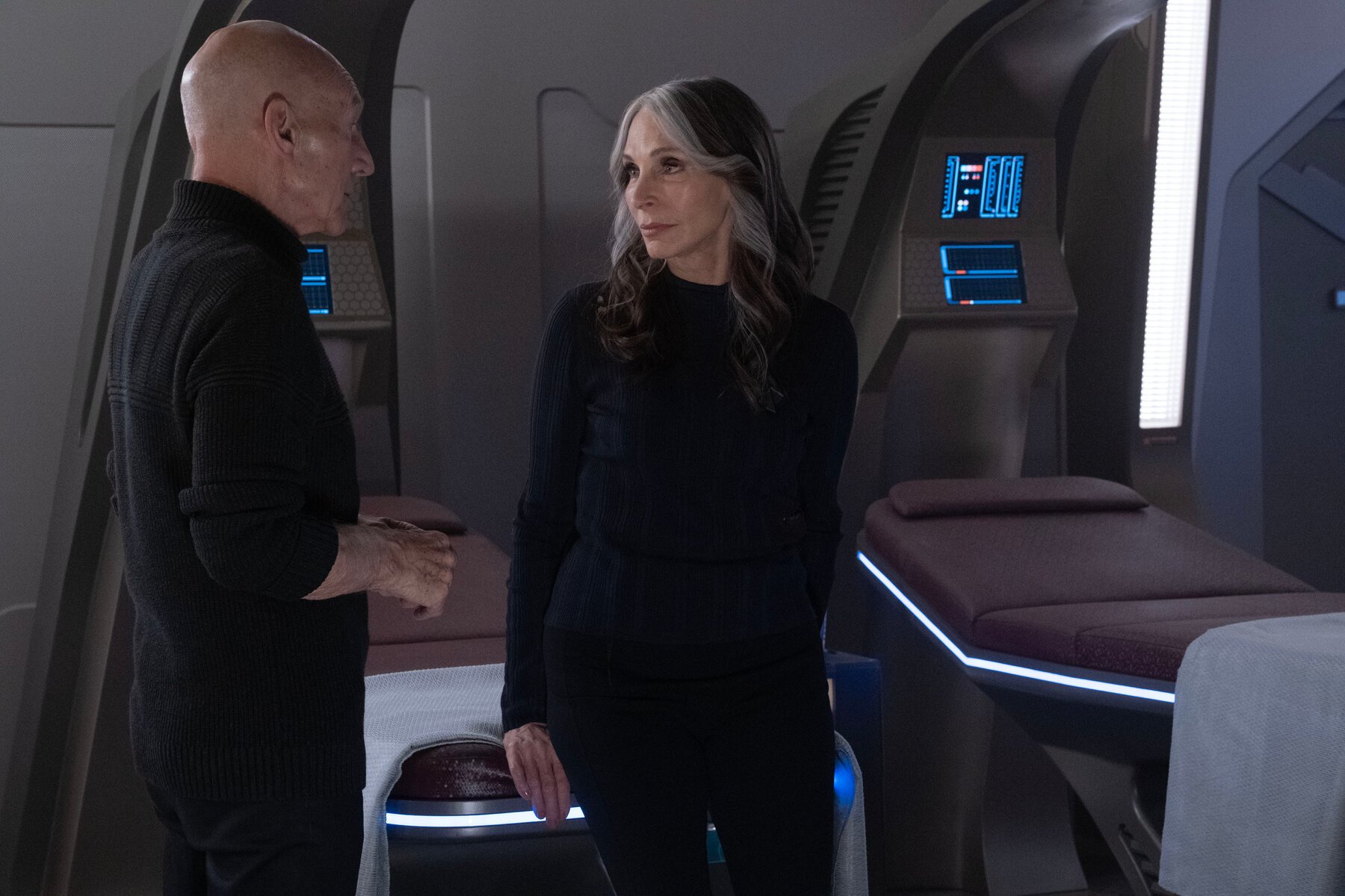 Patrick Stewart as Jean-luc Picard and Gates McFadden as Beverly Crusher in 'Star Trek: Picard.' They are in sickbay, and we see both of them from the thighs up. Picard is in profile as he talks to her. He is dressed in all black, and is bald. Beverly is leaning against a sickbay bed with her body turned toward the camera, but her head turned toward Picard. She's also dressed all in black. She has long, salt-and-pepper hair. 