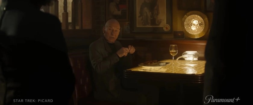 Image of Patrick Stewart as Jean-luc Picard on 'Star Trek: Picard.' He is seated in a booth at a bar with a plate of food and a glass of wine in front of him. It's a dark room, but there's a bright light at the table. He is trying to put his napkin in his lap as he looks up at someone talking to him. He's wearing a dark grey blazer and is bald. 