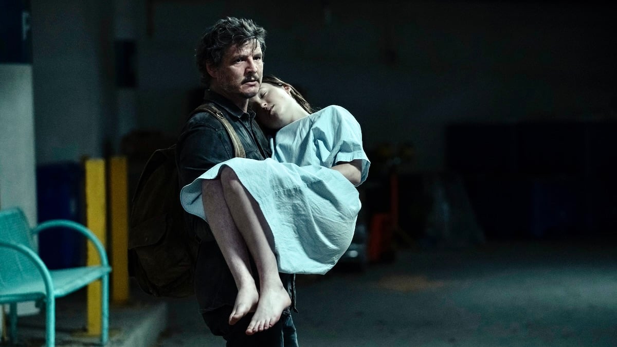 Image of Pedro Pascal (Joel) and Bella Ramsey (Ellie) in a scene from 'The last of us' on HBO. Joel has dark, scruffy hair and a mustache and wears a dark jacket and a brown backpack. He's carrying Ellie, who is unconscious and in a hospital gown. He's walking through a parking garage.