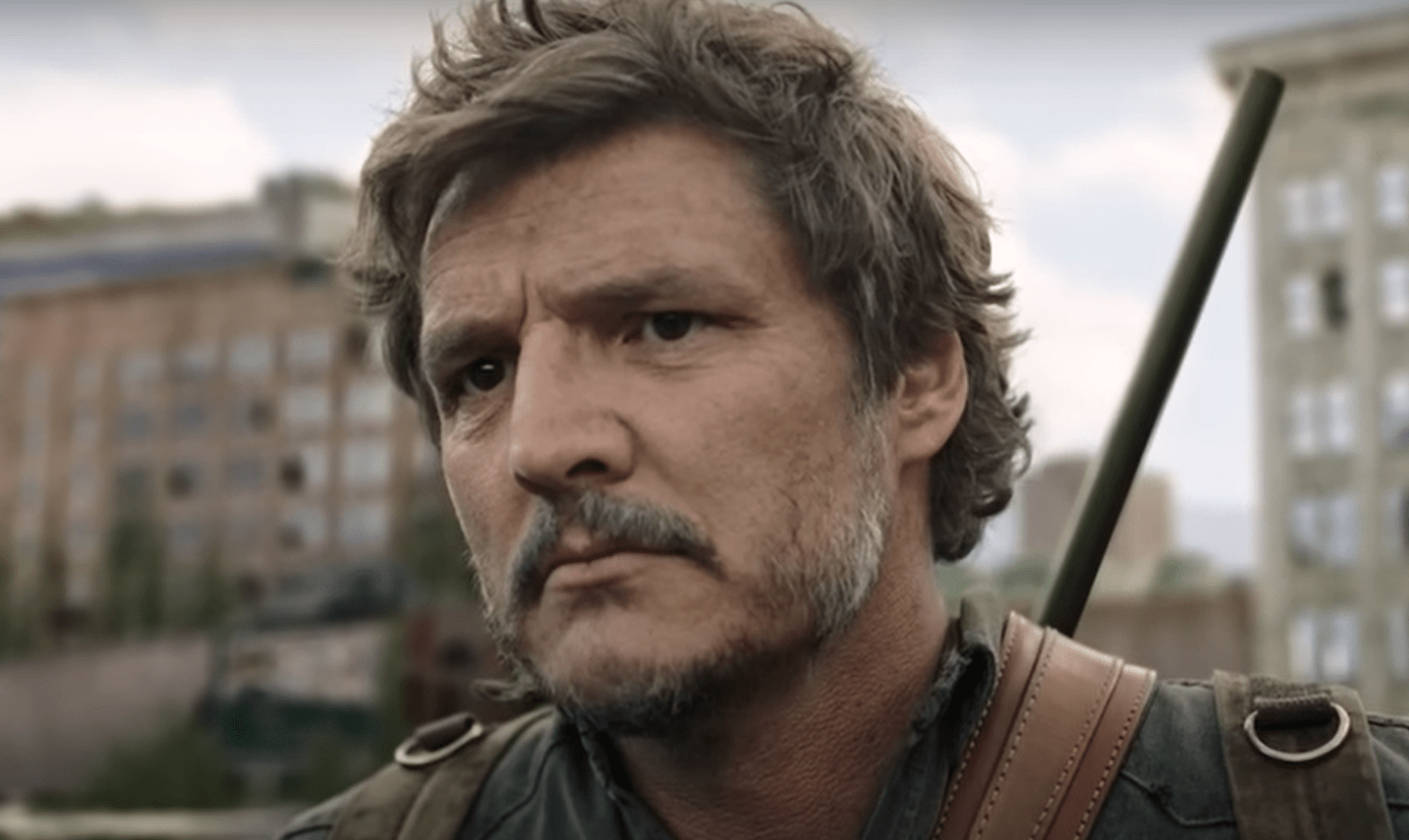 Image of Pedro Pascal as Joel on 'The last of Us' on HBO. Close-up image of Joel, who is outdoors and looking at something with a serious expression. His brown hair is shaggy, and he has a salt-and-pepper mustache and a light beard. He's wearing a rifle on his back and a green backpack and a greenish jacket. 