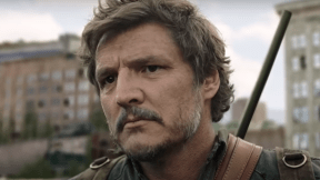 Image of Pedro Pascal as Joel on 'The last of Us' on HBO. Close-up image of Joel, who is outdoors and looking at something with a serious expression. His brown hair is shaggy, and he has a salt-and-pepper mustache and a light beard. He's wearing a rifle on his back and a green backpack and a greenish jacket.
