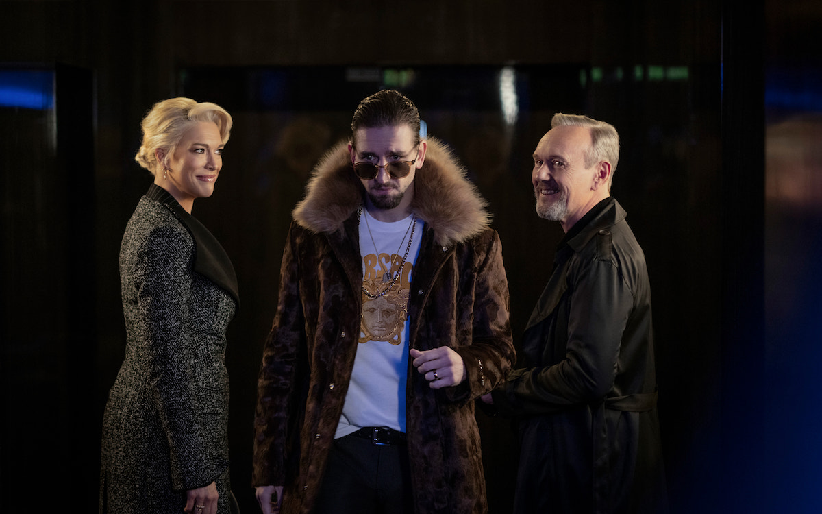 Rebecca, Zava, and Rupert stand in a hallway, smiling at each other tensely.