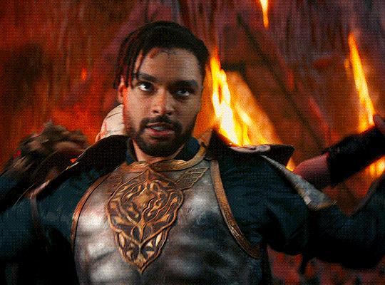 GIF of Regé-Jean Page as Xenk in 'Dungeons & Dragons: Honor Among Thieves.' As Xenk stands facing forward in a fiery underground lair, he reaches behind him with his sword to slit the throat of an undead attacker behind him. 
